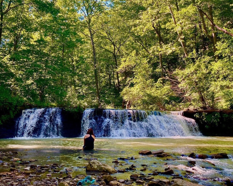 color photograph of a seated figure in shallow water, facing a waterfall, surrounded by trees