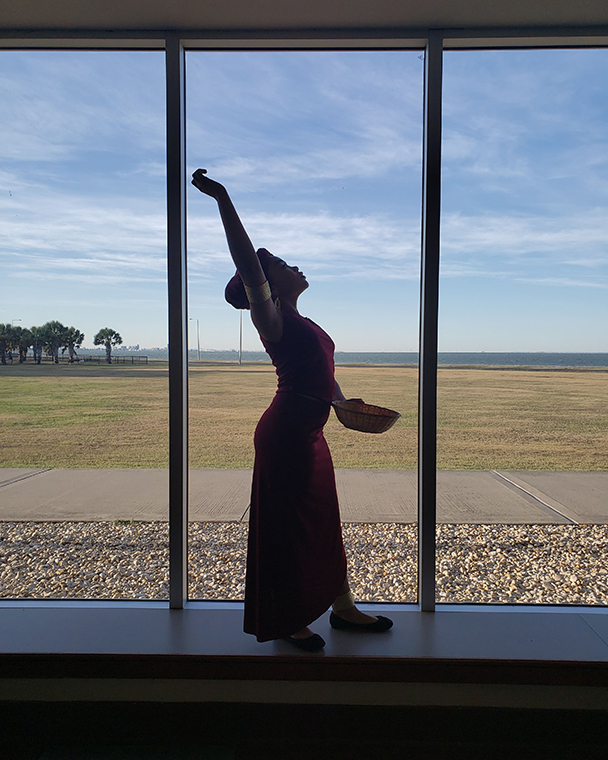 Color photograph of a young person in silhouette standing in front of large windows. The figure is looking up with one arm raised. The landscape outside the windows includes a sidewalk, lawn, palm trees, and body of water.