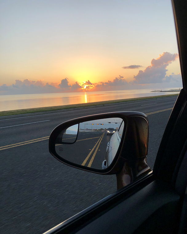 Color photograph of a cloudy sunrise taken through the driver's side window of an automobile. The sunrise is over the water and the side view mirror clearly reflects the road behind the car.