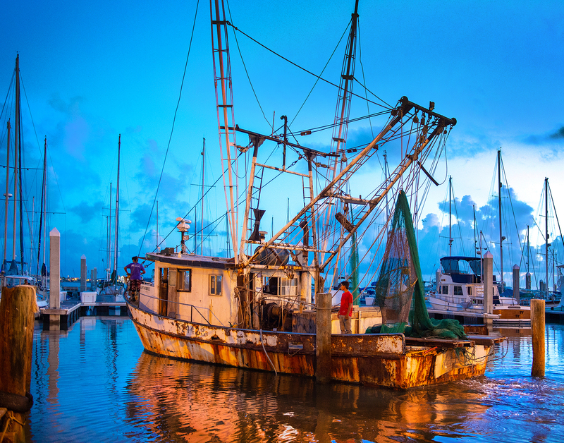 Colorful image of a shrimp boat in a harbor at sunrise. The boat is reflected on the water and the horizon is cloudy.