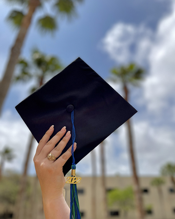 Color photograph of a hand holding a mortarboard up into the sky. The blue and green tassel is dated 2022, the hand holding the mortarboard is wearing a class ring. The background of the image includes palm trees, buildings, blue sky, and clouds.