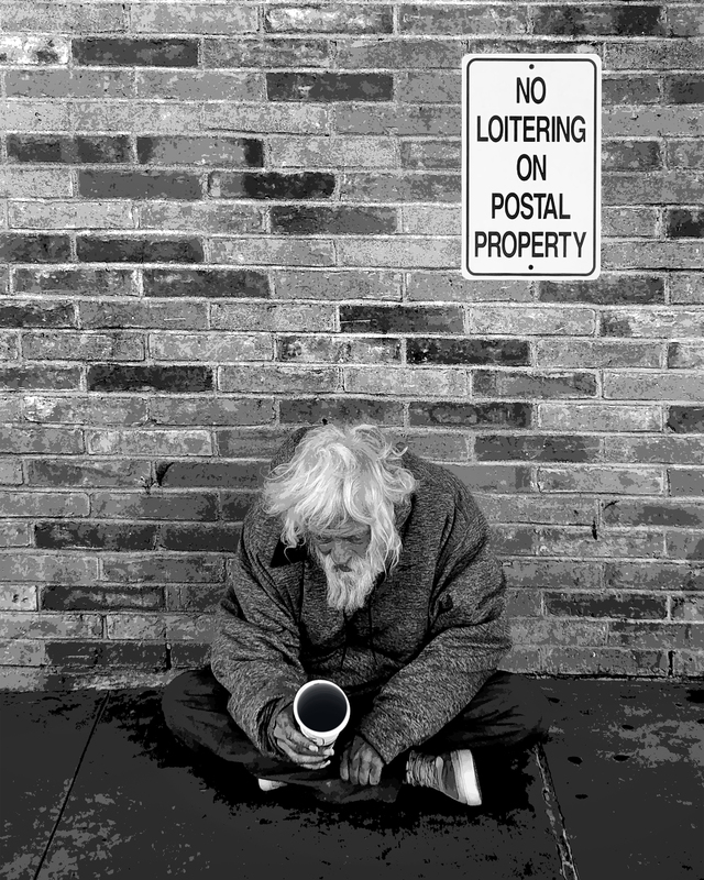 monotone photograph of a hunched figure seated on the ground beneath a no loitering sign posted on a brick wall