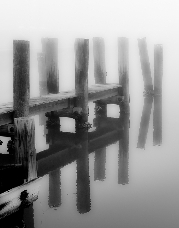 Black and white image of a pier in the fog. The pier is reflected in the water's surface.