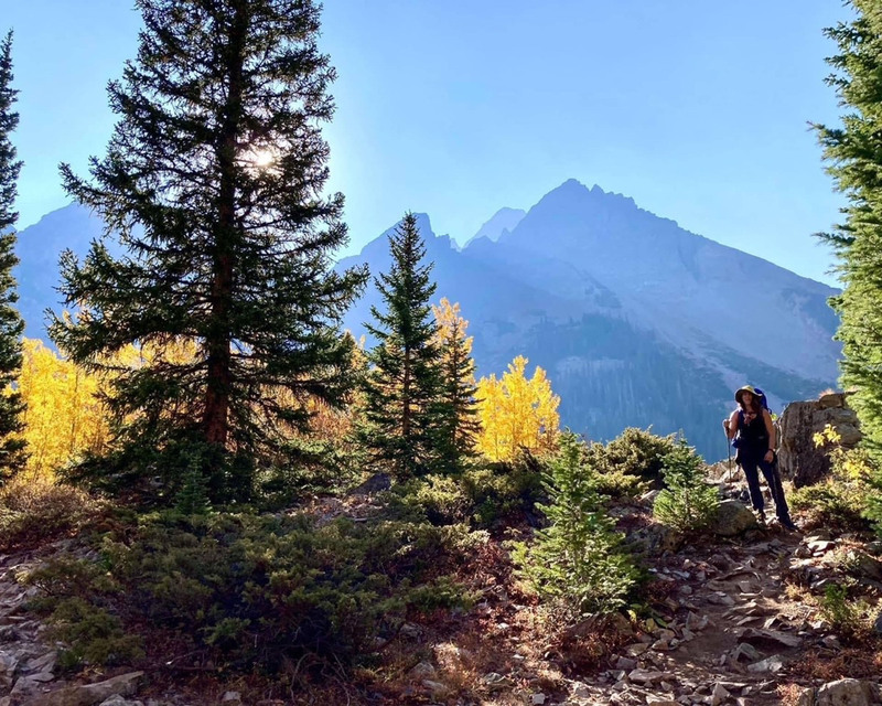 color photograph of a hiker on a rocky, tree lined trail, with mountains in the background