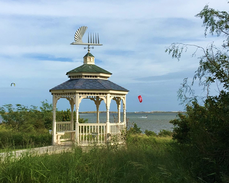 Color photograph of a gazebo on a grassy shoreline. Colorful kite surfers can be seen on the water.