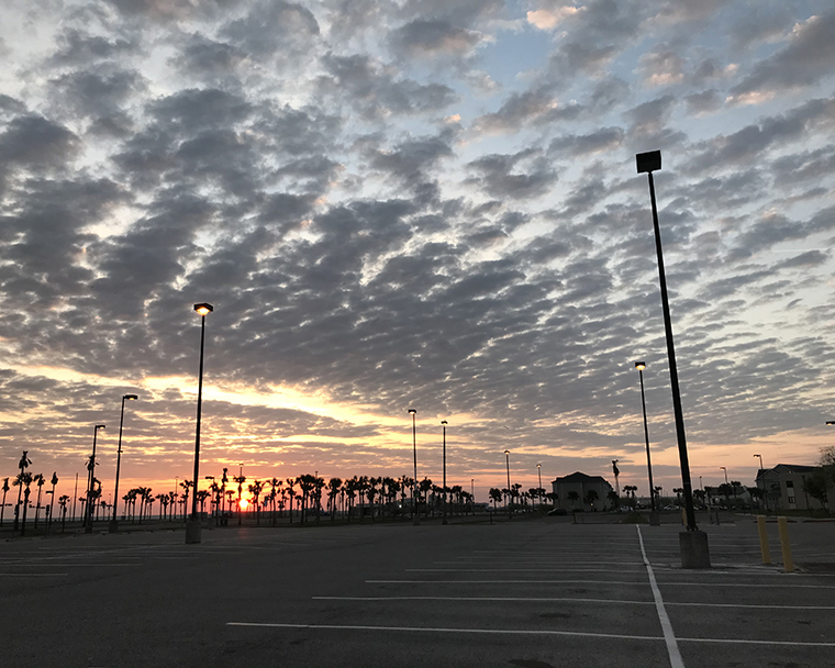 Color photograph of a sunrise through light clouds. The foreground includes an empty parking lot with palm trees in the distance.