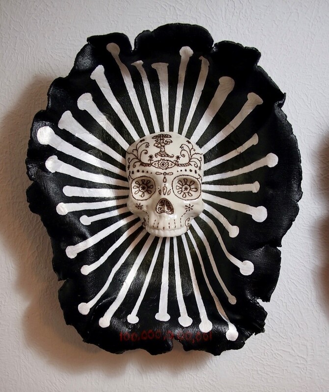 Ceramic art piece in an elongated bowl shape with rough edges. The bowl is black with white lines radiating from the center. The center has a skull figure, decorated in detail, using fire ants.