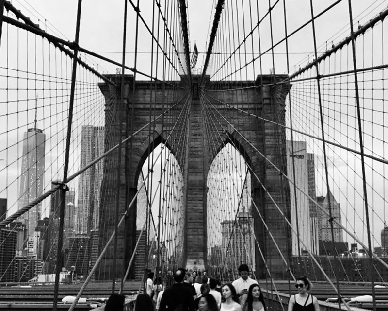 monotone photograph of pedestrians crossing a suspension bridge with a city skyline in the background