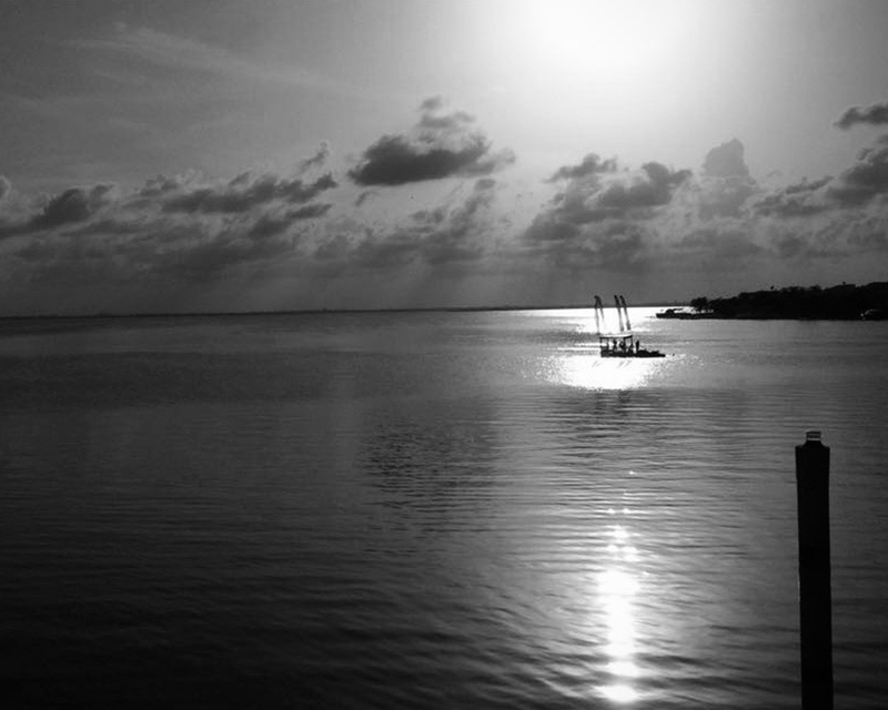 monotone photograph of a single boat, caught in the sun's reflection on the water near the shore.