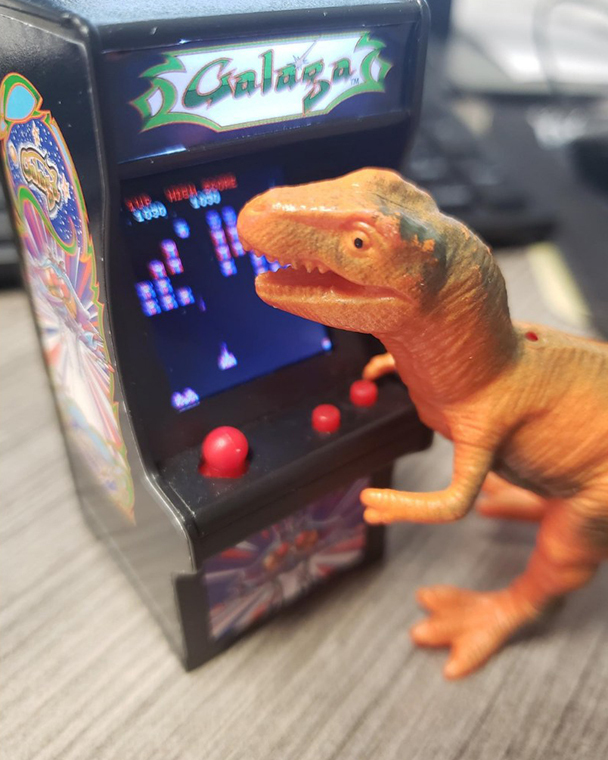 Color photograph of an orange plastic dinosaur posed in front of a miniature replica of a Galaga arcade game