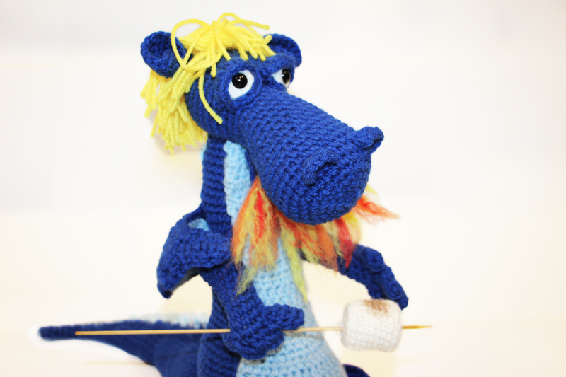 Crochet figure of a blue dragon. The dragon has yellow hair and is breathing fire to roast a marshmallow on a stick.