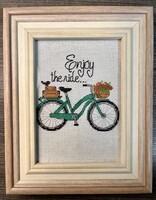Framed cross stitch of a green bicycle with a flowered basket on the handlebars. The words "Enjoy the Ride..." are stitched above the bicycle.